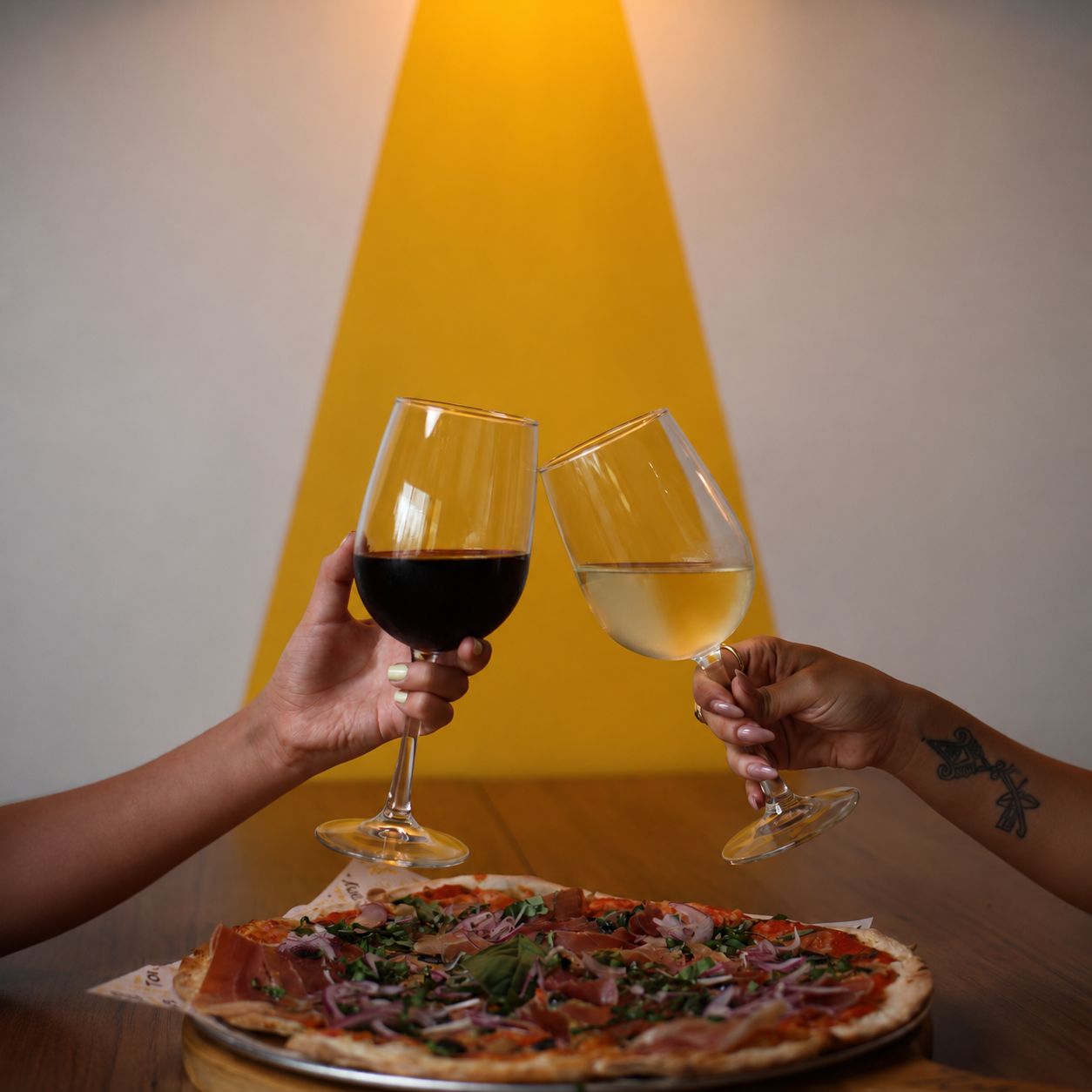 Dos manos brindando con copas de vino tinto y blanco sobre una pizza, con fondo amarillo